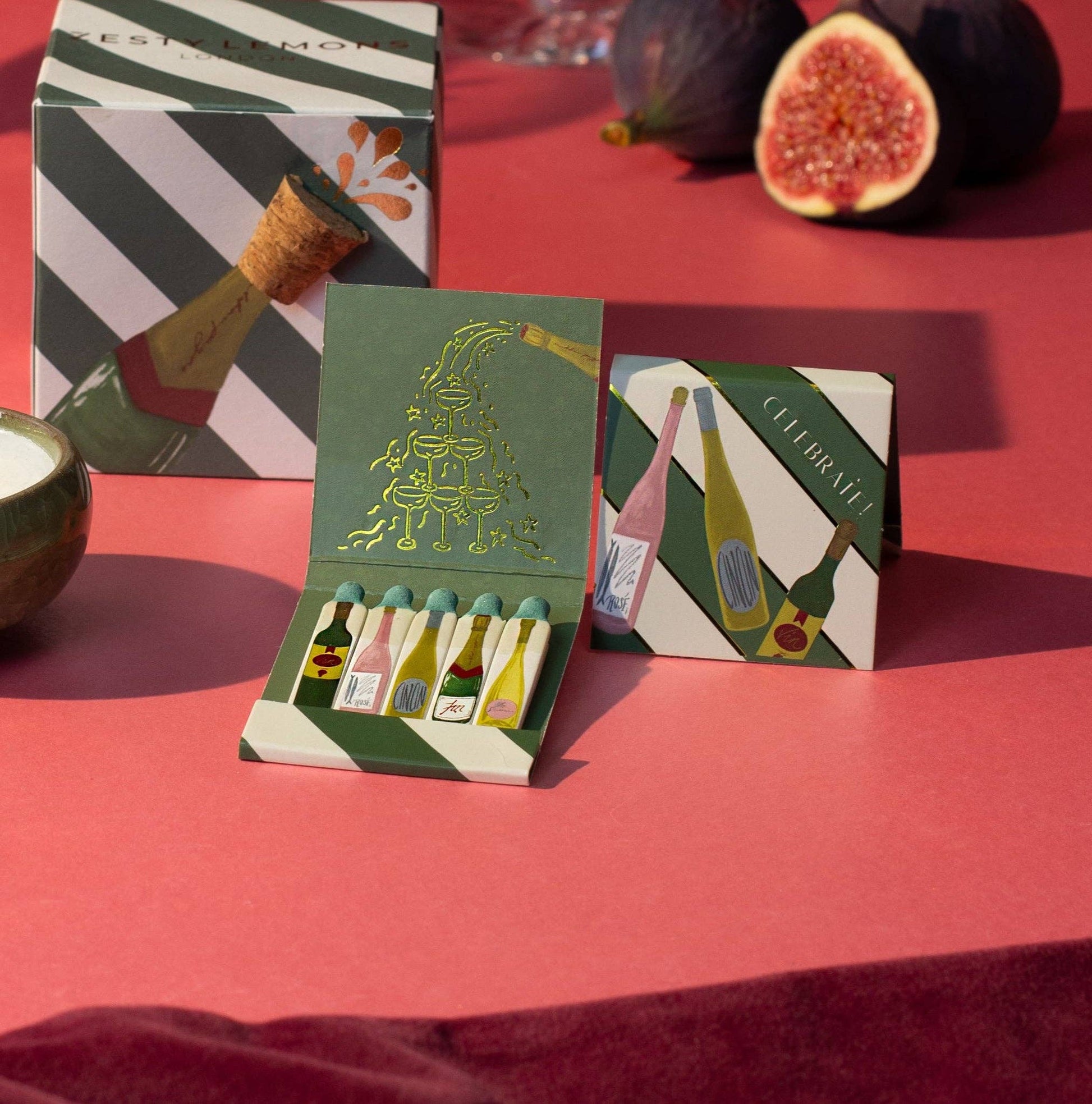 Boîte d’allumettes CELEBRATE de Zesty Lemons, illustrée de bouteilles festives avec allumettes décorées et intérieur doré.